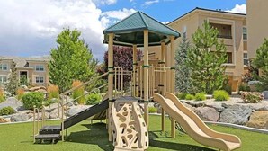 Un parque infantil con tobogán y estructura de escalada, rodeado de zonas ajardinadas y edificios residenciales.