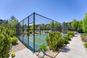 En tennisbana omgiven av ett staket, med en basketkorg, och med välskött grönska runt omkring. I bakgrunden syns bostadshus.