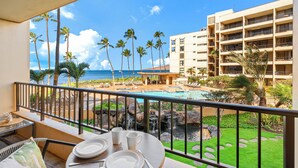 A balcony with a dining setup overlooking a pool area, palm trees, and a view of the ocean. - Gorgeous Oceanfront Remodel I Sugar Beach 210 (Kihei)