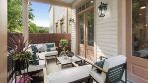 En overdækket terrasse med udendørs møbler, herunder en sofa og stole, omgivet af planter og et træhegn.
