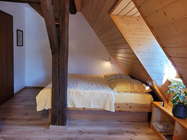 A bedroom with a sloped ceiling, a bed with a yellow blanket, a wooden shelf with a plant, and a framed picture on the wall.