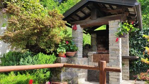 A rustic outdoor structure with stone walls and a wooden roof, surrounded by lush greenery and potted plants. - The mountain Cabin (scopa)