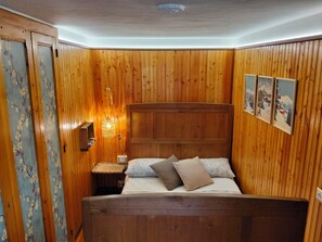A wooden-paneled room with a bed, a nightstand, a wall-mounted lamp, and framed pictures. - The mountain Cabin (scopa)