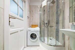 Un baño pequeño con lavadora, cabina de ducha con mampara de vidrio y lavamanos.