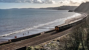 A coastal railway track with a train, a beach, and a distant hill.