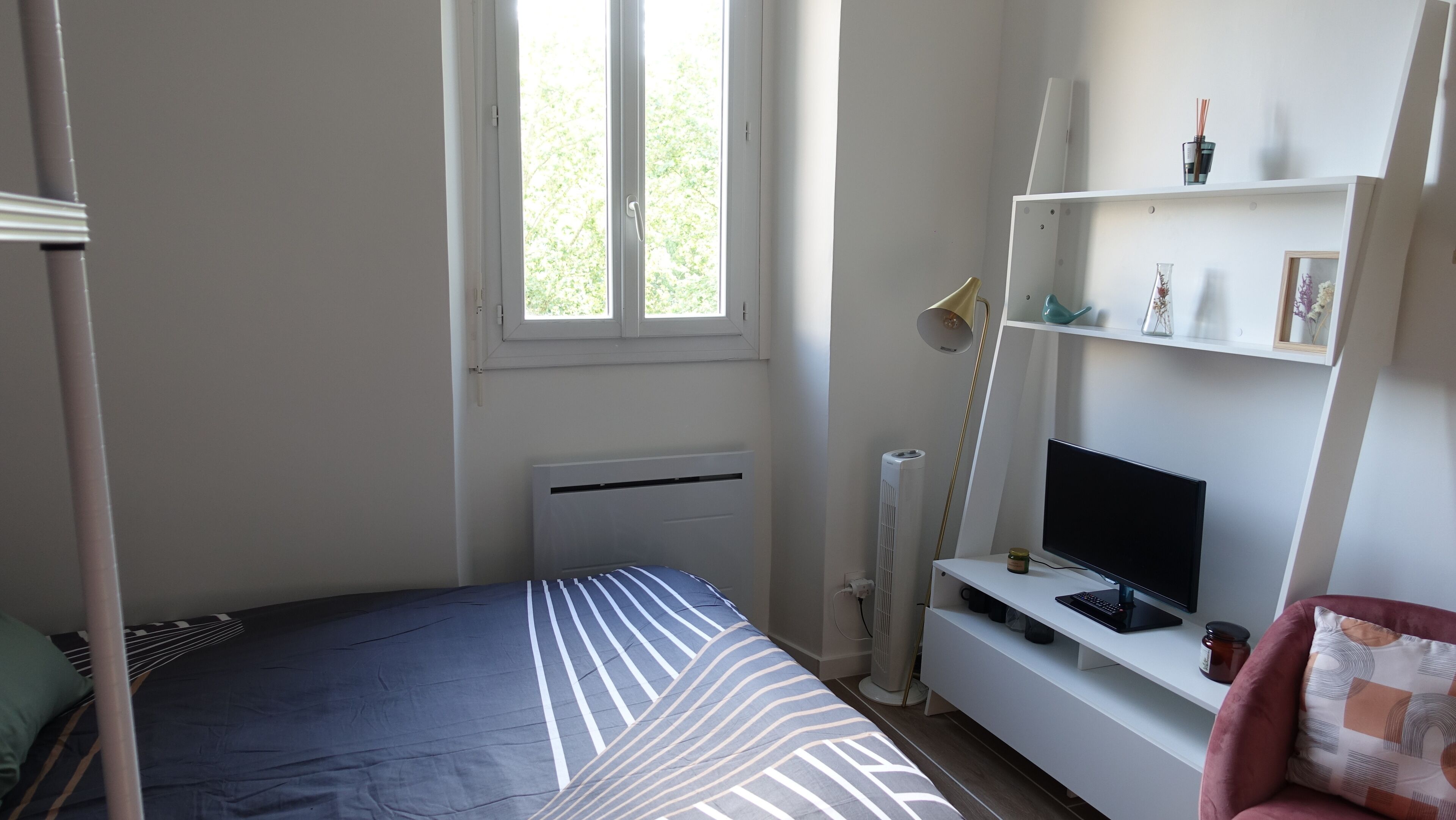 A bedroom with a bed, a TV, a shelf with decorative items, and a window.