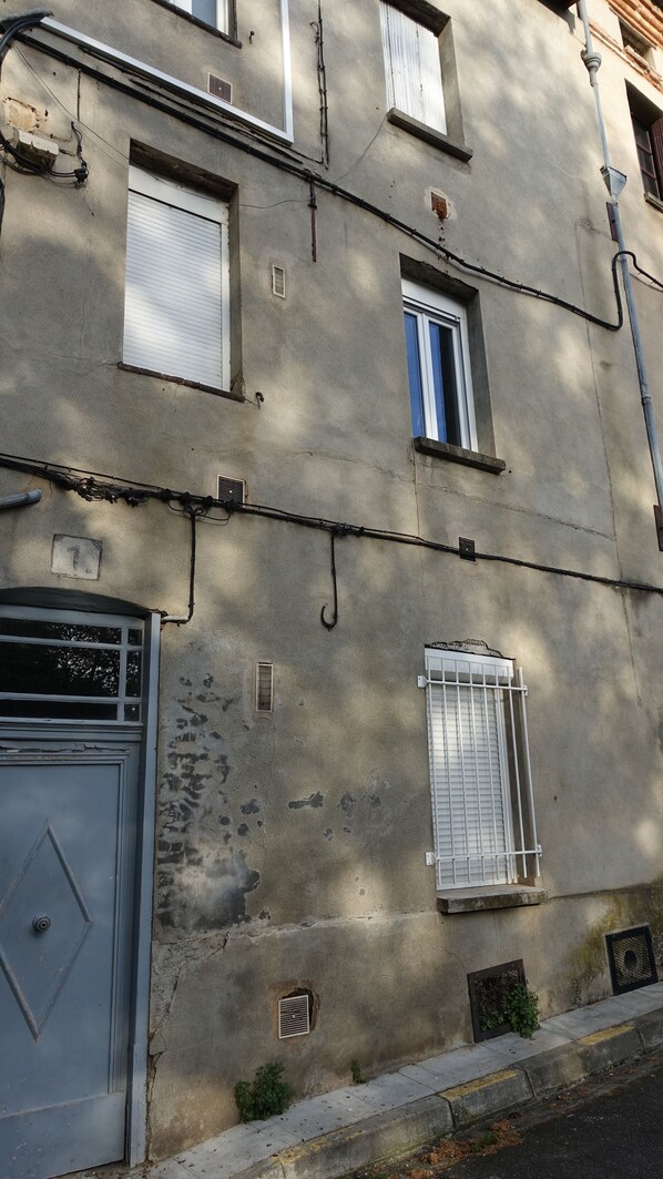 Un bâtiment doté de nombreuses fenêtres, dont certaines sont équipées de volets, et d’une porte ornée de motifs géométriques. On peut observer des fissures et des zones réparées sur les murs, et une petite plante pousse près de la base du bâtiment.