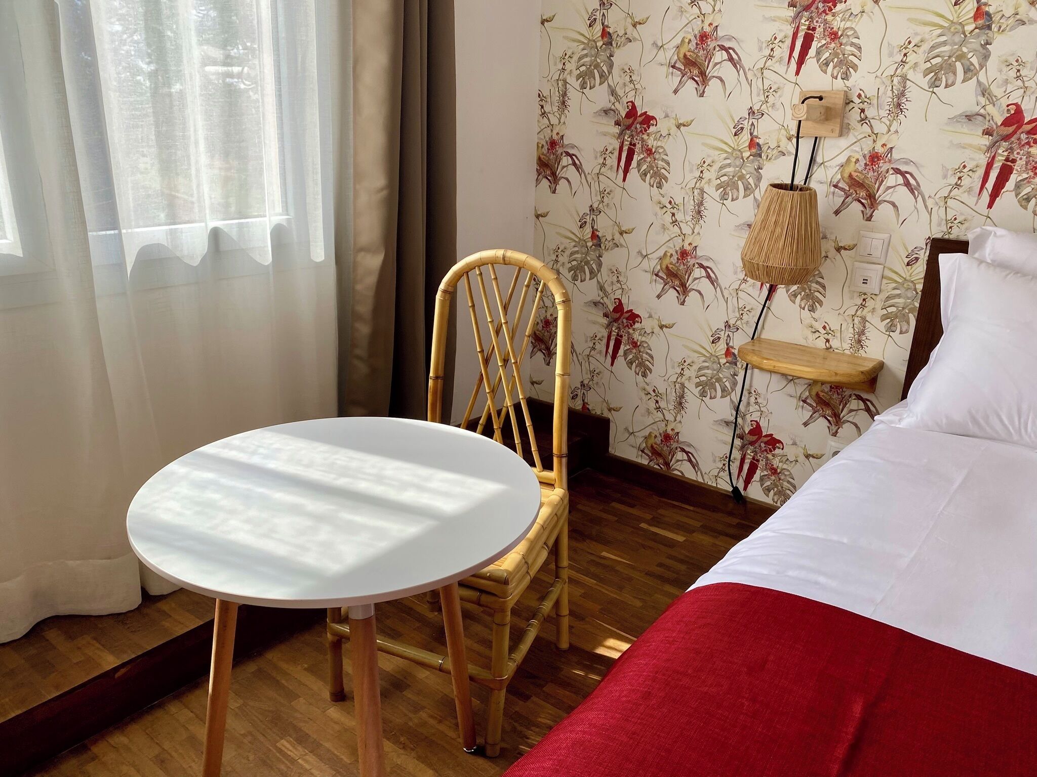 Une pièce comprenant un lit, une table ronde, une chaise en osier et un mur recouvert d’un papier peint à motifs floraux.