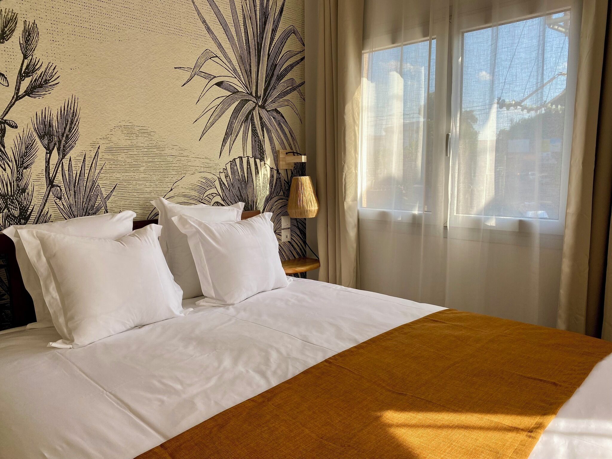 Habitación de hotel con una cama grande, ropa de cama blanca, un muro cubierto de plantas tropicales, una ventana con cortinas translúcidas y una mesita de noche de madera con una lámpara.