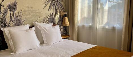 Habitación de hotel con una cama grande, ropa de cama blanca, un muro cubierto de plantas tropicales, una ventana con cortinas translúcidas y una mesita de noche de madera con una lámpara.