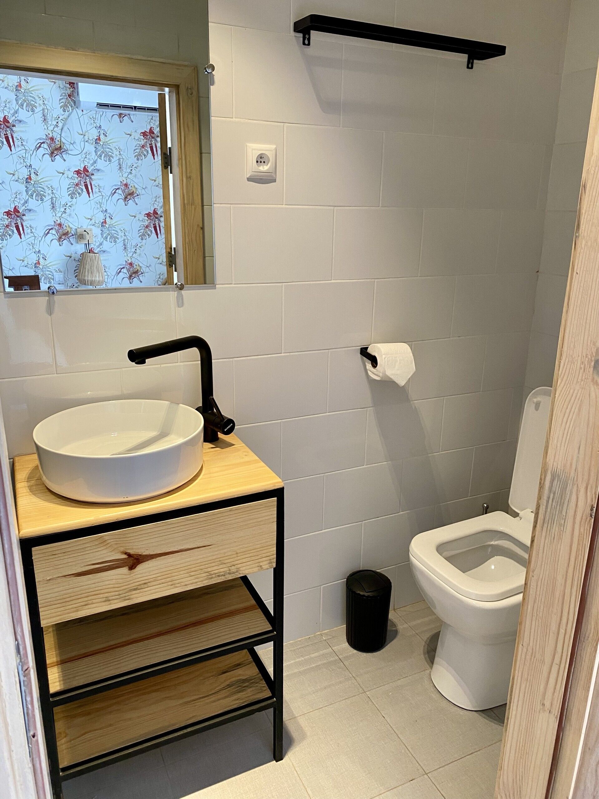 A bathroom with a white sink, black faucet, and a wooden vanity. There is a toilet and a black trash bin.