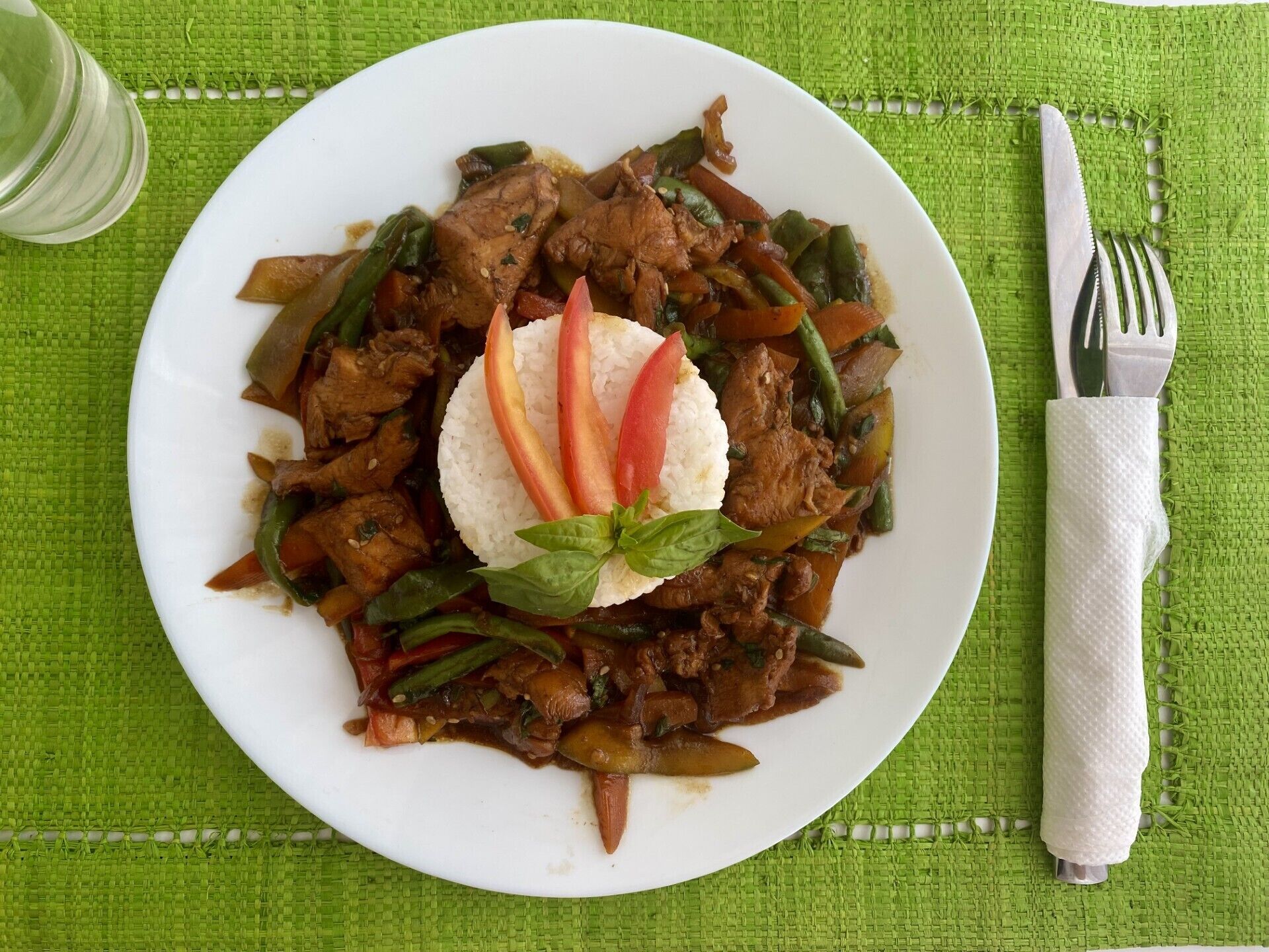 Un plato de carne y vegetales salteados, acompañado de arroz blanco, adornado con albahaca y rodajas de chile rojo, sobre una base de mimbre verde.