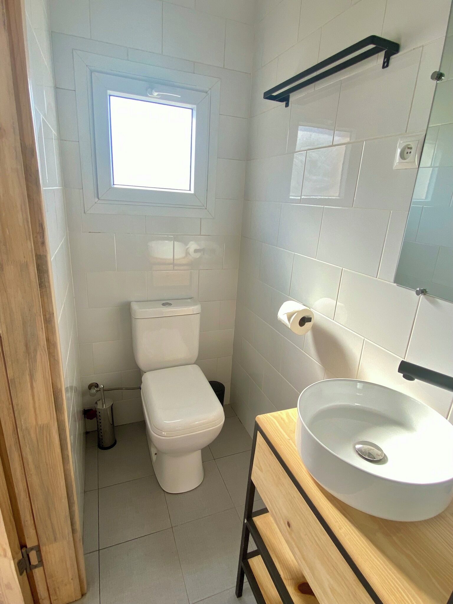 A small, clean bathroom with white tiles, a toilet, a sink, and a window providing natural light.