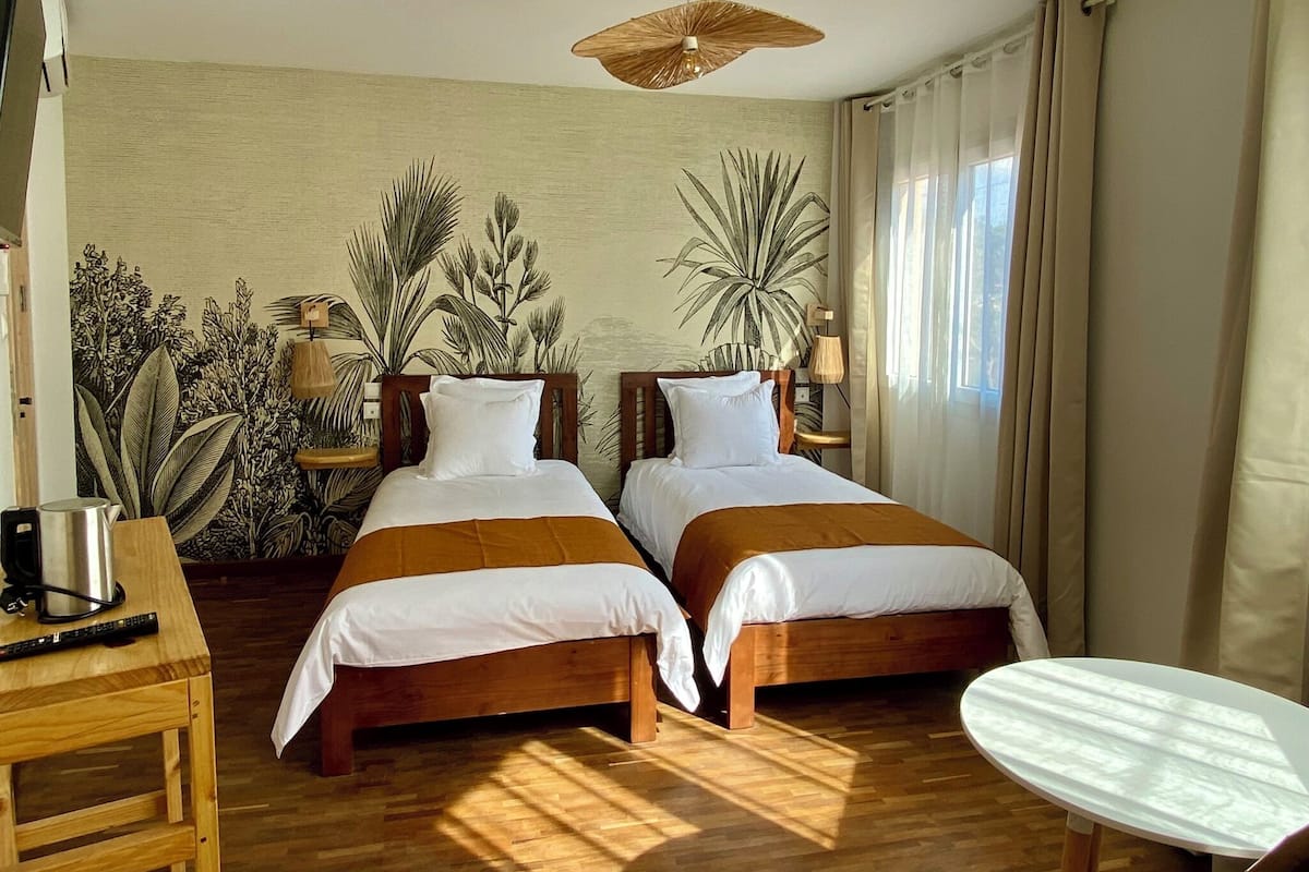 A hotel room with two wooden beds, a small table, a coffee maker, and a large window with curtains.