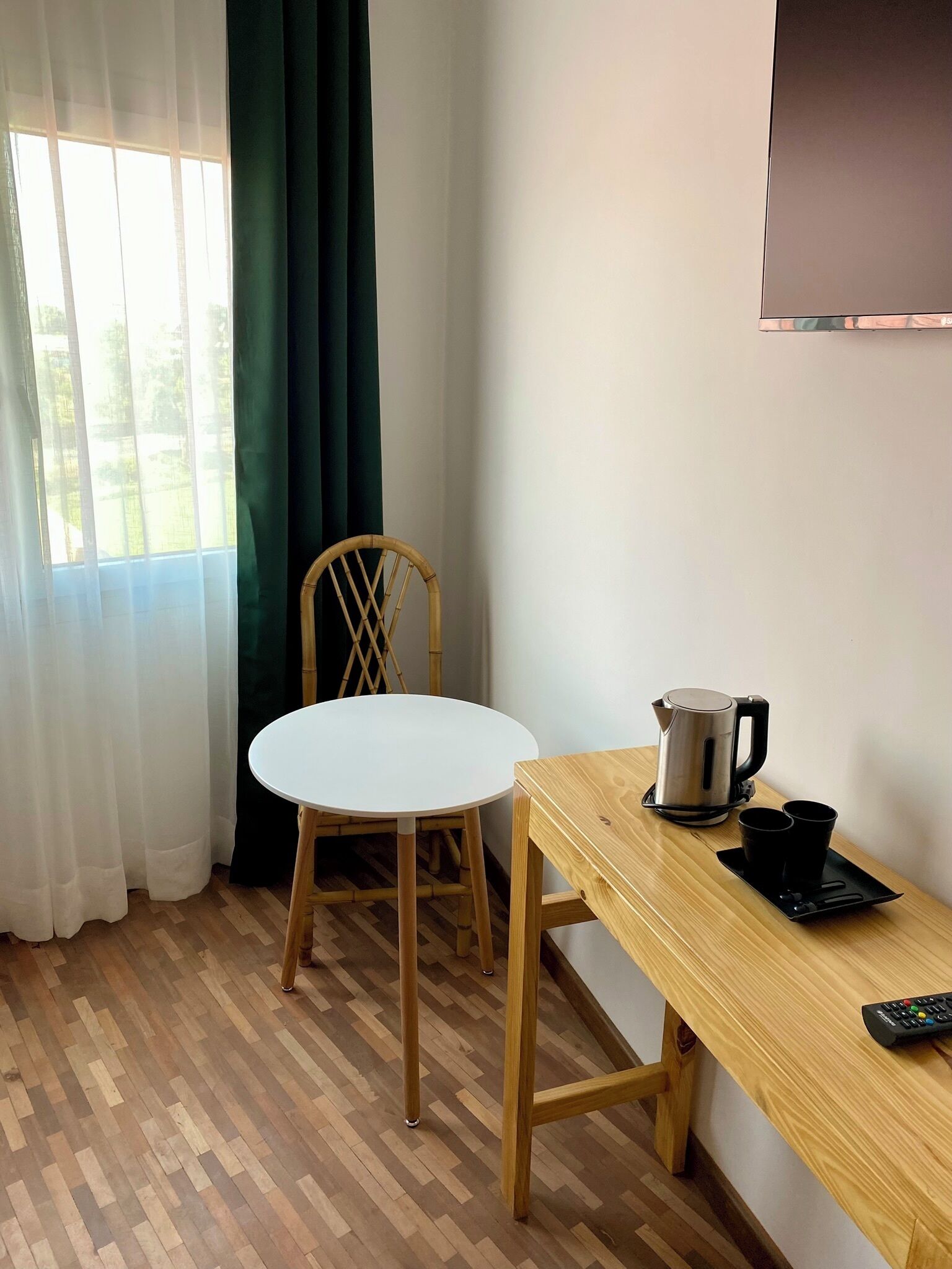 Una habitación con piso de madera, una mesa blanca, una silla de bambú, una cafetera y un control remoto.