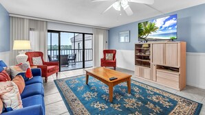 A living room with a blue sofa, red armchairs, a wooden coffee table, and a TV on a wooden stand. - Pelican's Perch - Expansive Condo with Water Views, Pool-Across from Beach (Indian Shores)