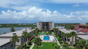 A hotel with a central courtyard featuring a swimming pool, surrounded by palm trees and a view of a body of water. - Pelican's Perch - Expansive Condo with Water Views, Pool-Across from Beach (Indian Shores)