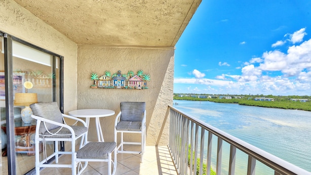A balcony with a view of a river, white wicker chairs, a small table, and a decorative wall with palm trees. - Manatee Zone,SeaClub #405~Corner unit with incredible water front views (Indian Shores)