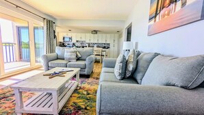 A living room with a grey sofa, a coffee table, and a colorful rug. There is a kitchen in the background with white cabinets and a dining table.