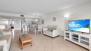 A modern living room with a white sofa, a wooden coffee table, a TV on a white stand, and a kitchen in the background. - Isle Be Back,SeaClub #402~Water View, Pool,Fishing (Indian Shores)
