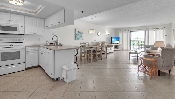A modern kitchen with white cabinetry, a central island, and a dining area with a table and chairs. - Isle Be Back,SeaClub #402~Water View, Pool,Fishing (Indian Shores)