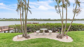 A landscaped area with a table and benches, palm trees, and a view of a lake with a dock and a building in the background. - Coral Reef Retreat, SeaClub #44~Pool, Fishing (Indian Shores)