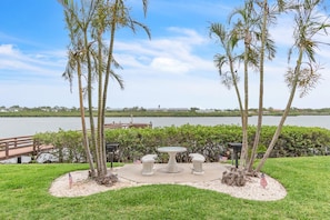 A landscaped area with a table and benches, palm trees, and a view of a lake with a dock and a building in the background.