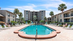 A swimming pool surrounded by lounge chairs and palm trees, with a multi-story building in the background. - Coral Reef Retreat, SeaClub #44~Pool, Fishing (Indian Shores)