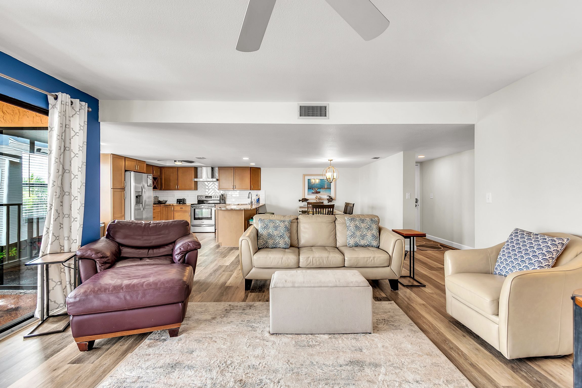 A modern living room with a leather sofa, a beige sofa, and a coffee table. There is a kitchen in the background with wooden cabinets and a dining area with a table and chairs.