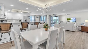 Eine moderne Küche mit weißen Schränken, ein Essbereich mit weißem Tisch und Stühlen sowie ein Wohnzimmer mit Sofa und Fernseher.