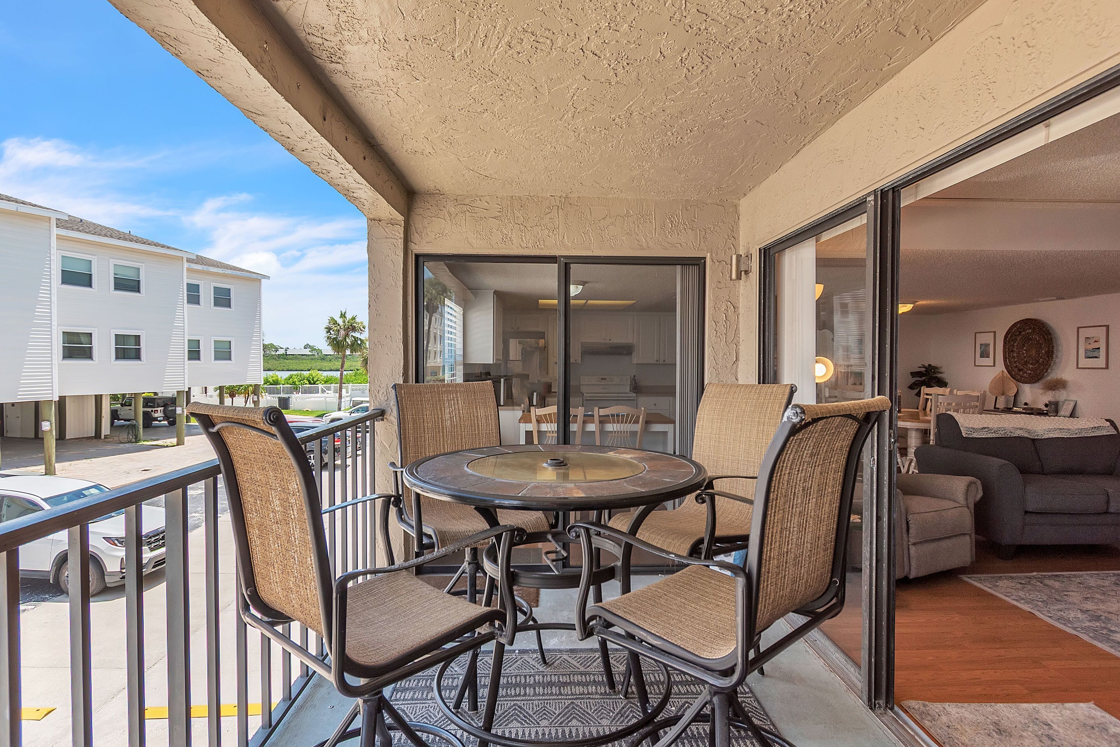 A balcony with a round table and chairs, a sliding glass door leading to a living area, and a view of a parking lot and buildings.