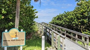 A wooden bridge leading to a beach access park with lush greenery and a clear blue sky.