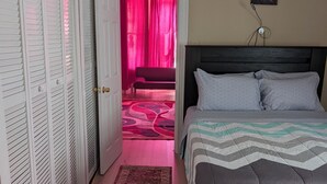 A bedroom with a bed, a wardrobe, a door leading to a sitting area, and a wall-mounted lamp.