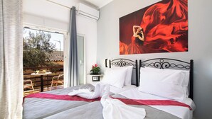 Una cama bien tendida con sábanas blancas y una manta roja, un cuadro grande de color rojo en la pared, y un balcón con mobiliario de exterior.