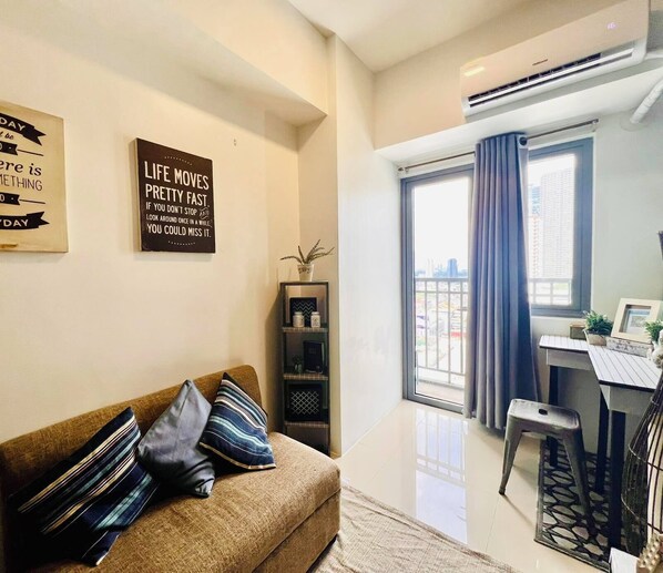 A modern living room with a sofa, a small side table, and a desk with a chair. There are two wall decorations and a window with curtains. - Ortigas Business District Residences (Mandaluyong)