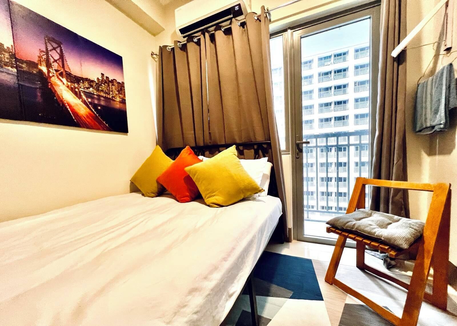 A hotel room with a bed, pillows, a chair, a window with curtains, and a painting on the wall.