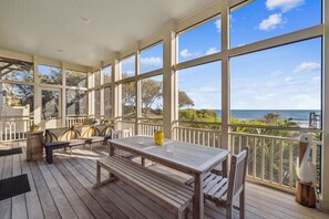 Une terrasse couverte avec une table et des chaises en bois, de grandes fenêtres offrant une vue sur l’océan.