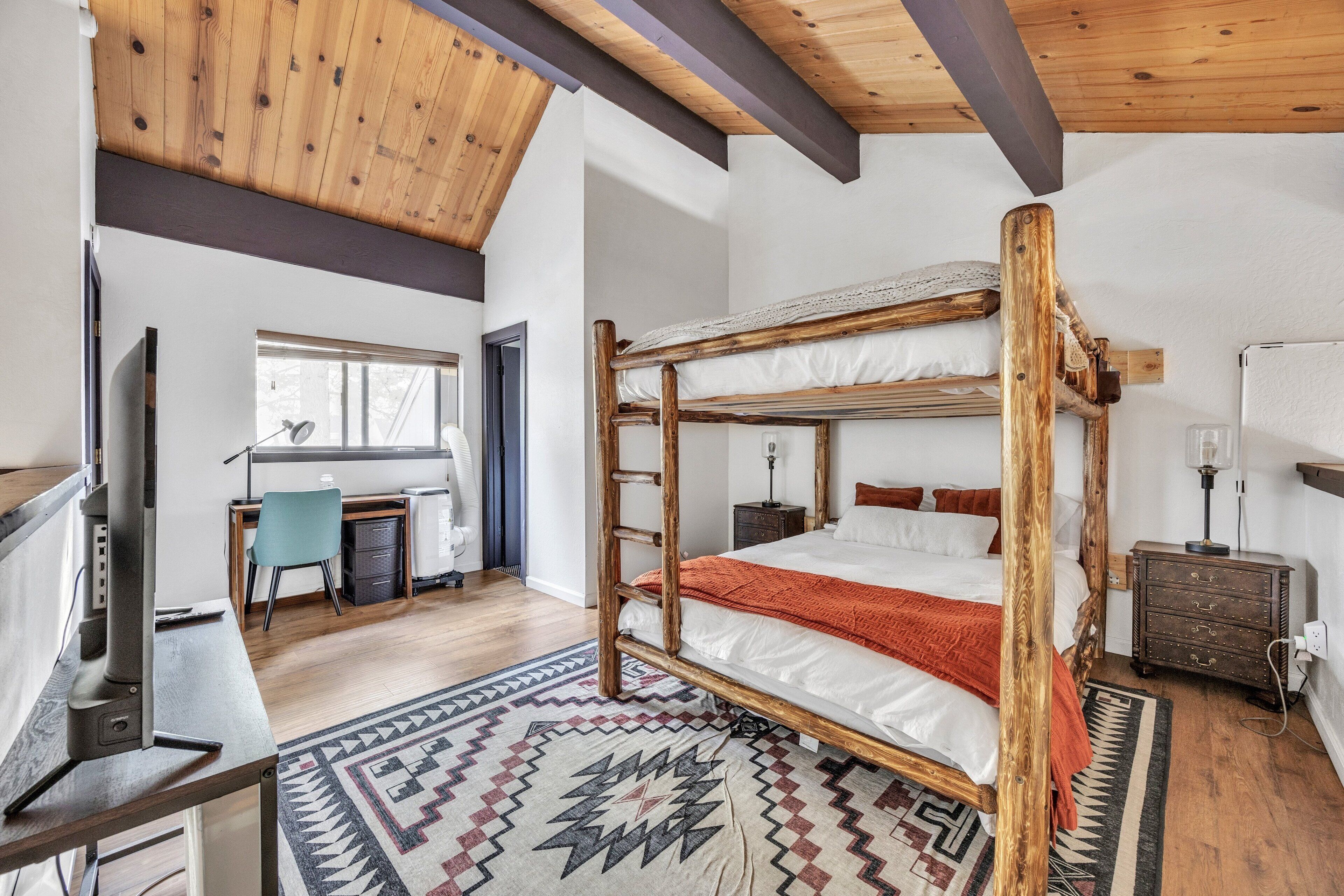 Een slaapkamer met een houten stapelbed, een bureau met stoel, een televisie en een kamerkleed met dessin.