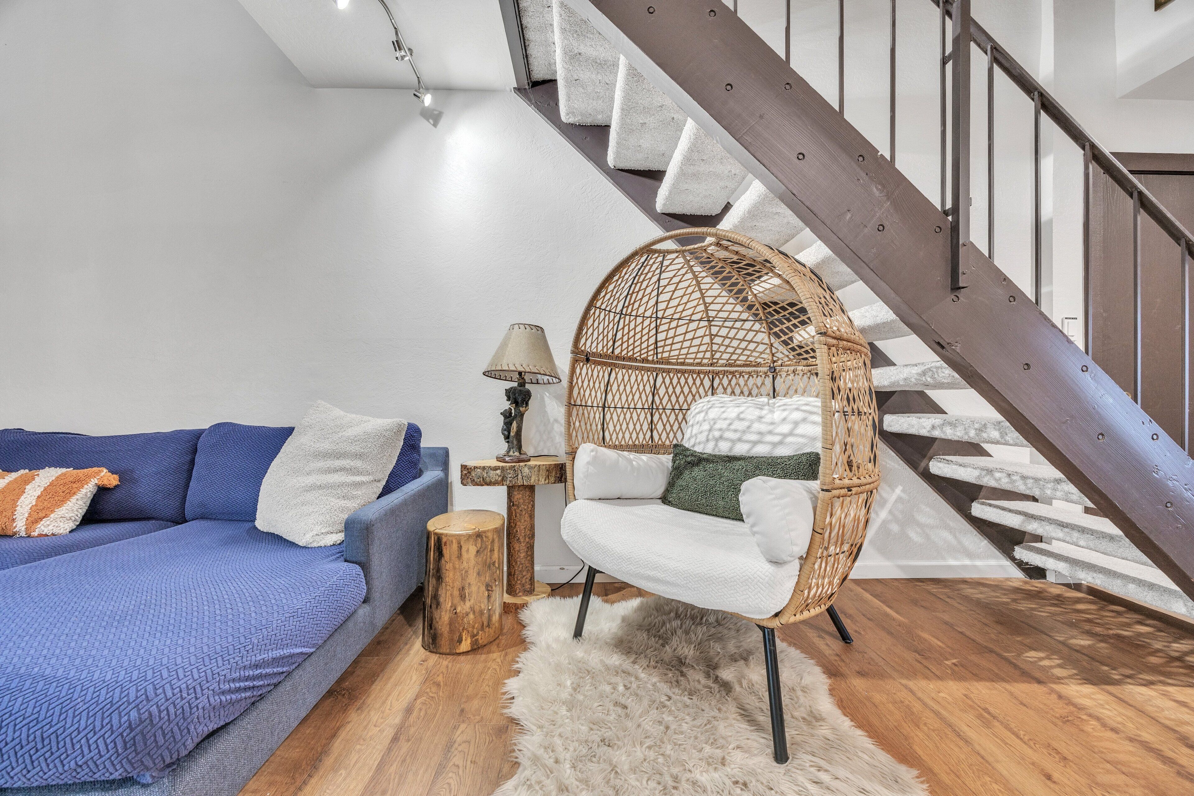 A modern living room with a blue sofa, a wicker chair, a small wooden table with a lamp, and a staircase.