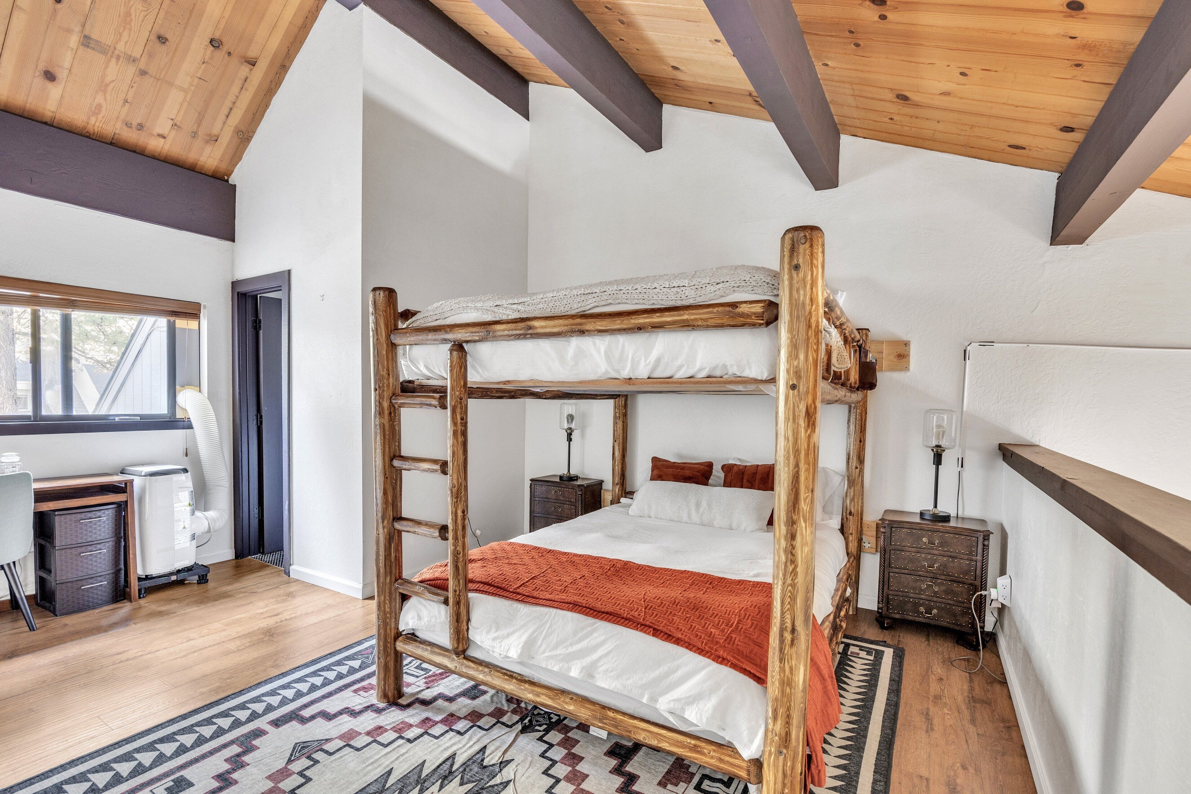 A rustic bedroom with a wooden bunk bed, a desk with a chair, a patterned rug, and a window with a view.