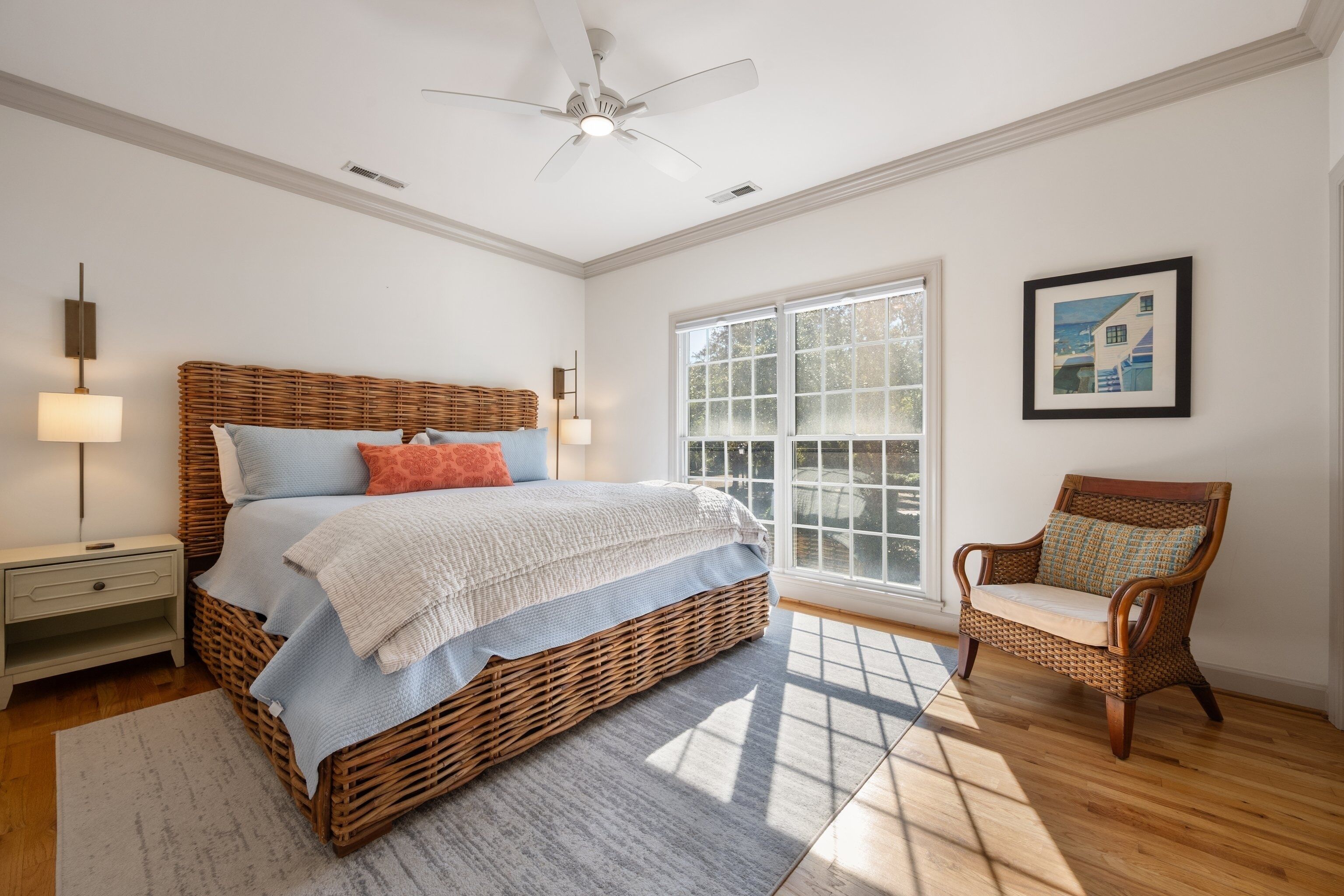 A bedroom with a large bed, a wicker chair, a window with blinds, and a ceiling fan.