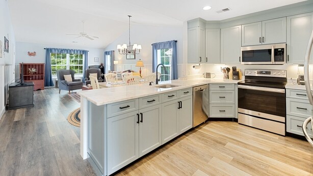 A modern kitchen with grey cabinets, a central island, stainless steel appliances, and a dining area with a grey sofa and blue curtains. - 65d Isle Awhile 3 Bedroom Condo (Pawleys Island)