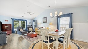 A spacious living room with a dining area, featuring a ceiling fan, a chandelier, and a large window with curtains. - 65d Isle Awhile 3 Bedroom Condo (Pawleys Island)