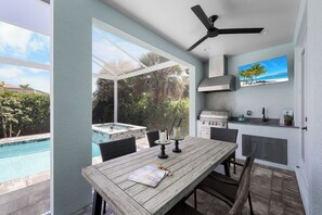 A patio with a wooden table, chairs, a grill, and a TV. - 2023 Custom Built Home | Heated Pool & Hot Tub (Marco Island)