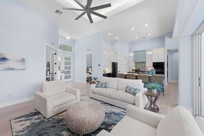 A spacious living area with a large sofa, a round ottoman, and a dining table. There is a kitchen in the background with modern appliances.