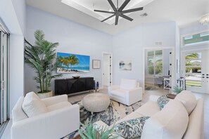 A spacious living room with a large ceiling fan, a white sofa set, a large potted plant, and a painting of a beach scene. - 2023 Custom Built Home | Heated Pool & Hot Tub (Marco Island)