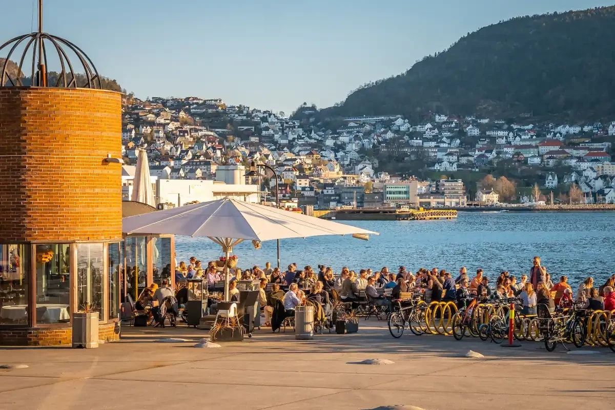 Ein Café am Wasser mit Außenbestuhlung, ein Backsteinhaus und Blick auf eine Hügelstadt.