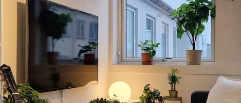 A cozy living room with a flat-screen TV, a guitar, a green side table with plants, a white sofa, and a window with a view of buildings.