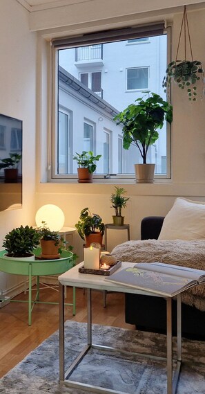 Una sala de estar acogedora con un ventanal grande, una mesa verde con plantas, una vela y un libro.