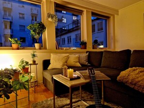Ein gemütliches Wohnzimmer mit einer Couch, einem Couchtisch und einer Gitarre. Es gibt Topfpflanzen und ein Fenster mit Blick auf Gebäude.